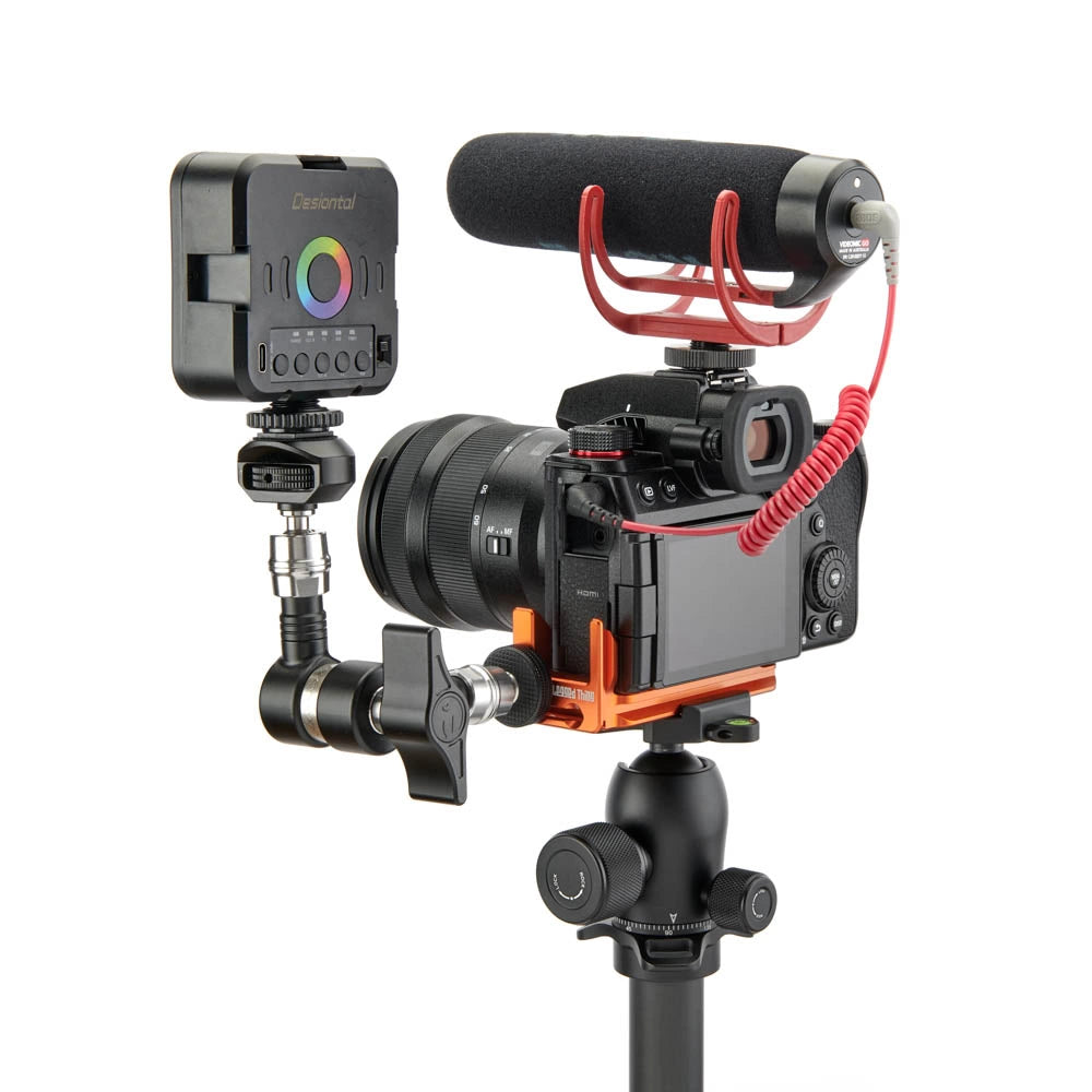 A camera rig setup featuring a black camera body with an orange anodized aluminum mount, a black shotgun microphone with a red coiled cable, a wireless transmitter, and a black tripod, all isolated on a white background.