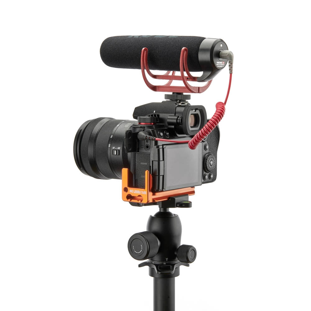 A camera rig setup featuring a black camera body with an attached lens, an orange quick-release bracket, a black tripod mount, a red coiled cable, and a black shotgun microphone on a red stand against a solid white background.