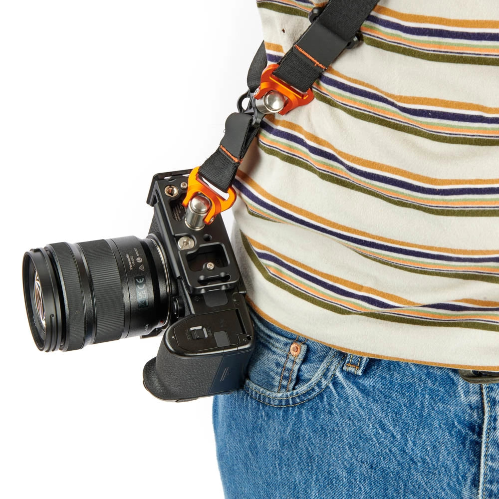 A close-up of a person wearing a striped shirt and blue jeans, carrying a black camera with an orange and black sling strap across their torso against a white background.