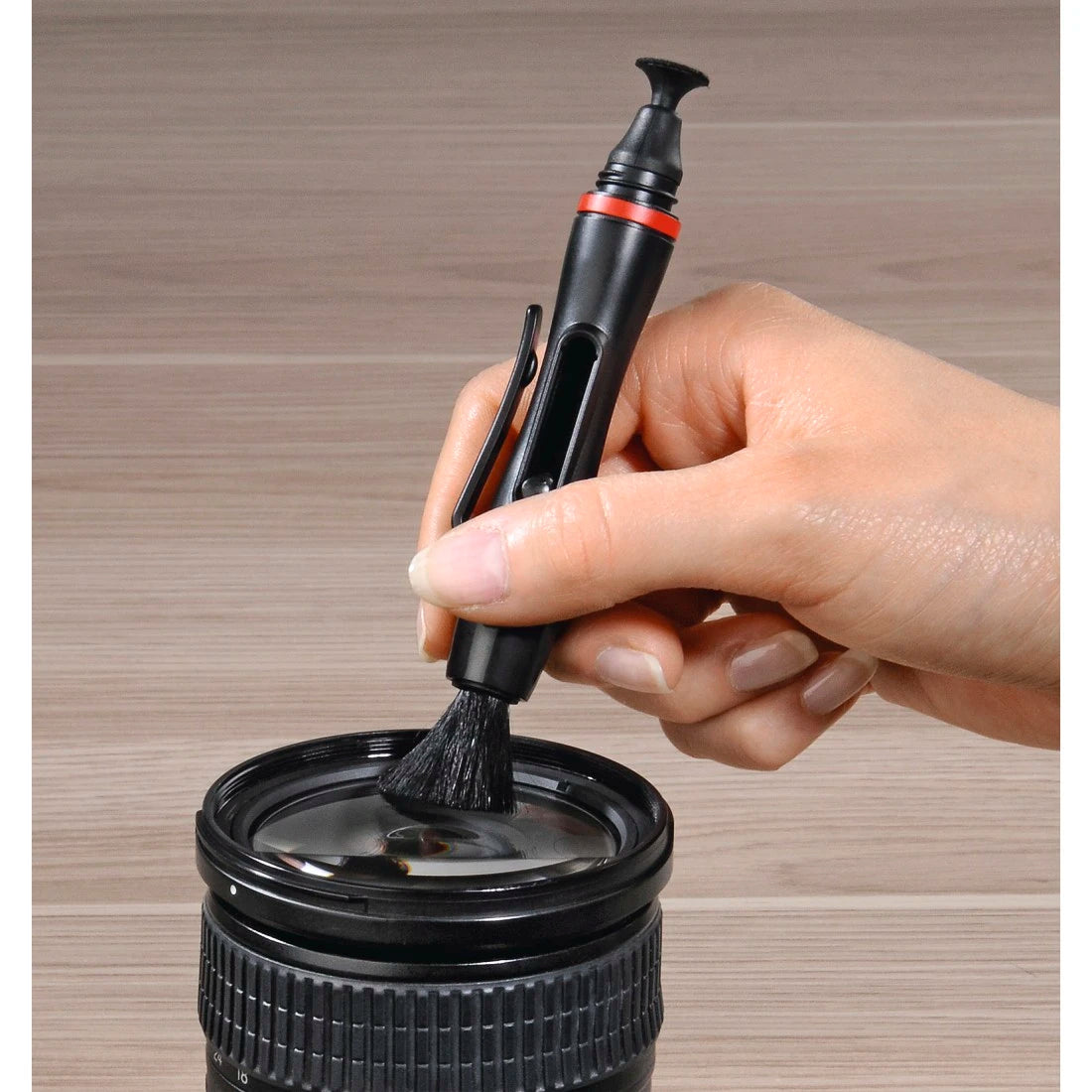 Hand using a lens cleaning brush on a camera lens against a wooden background