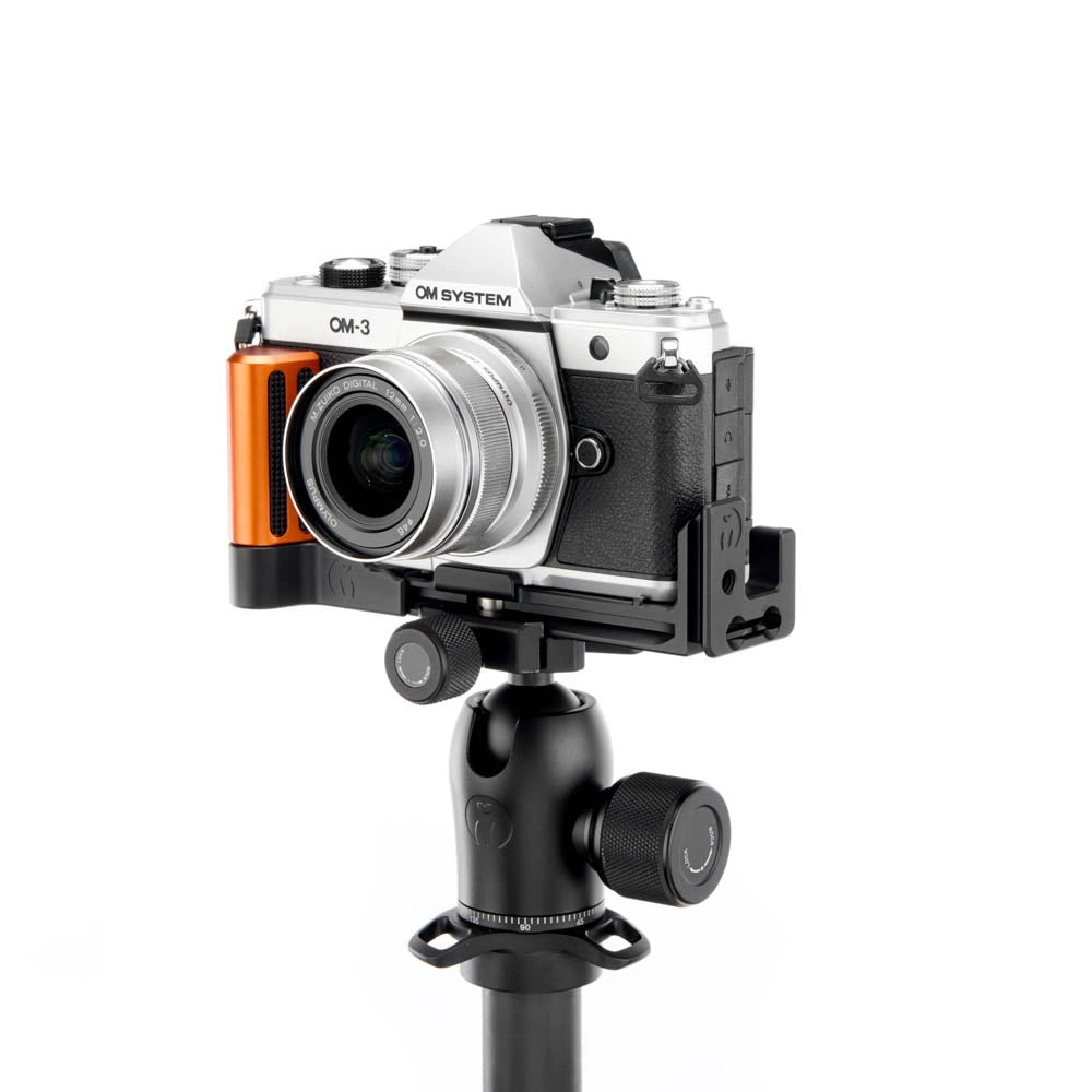 A silver and black OM-3 mirrorless camera with an orange accent grip, mounted on a black tripod mount, shown against a solid white background.