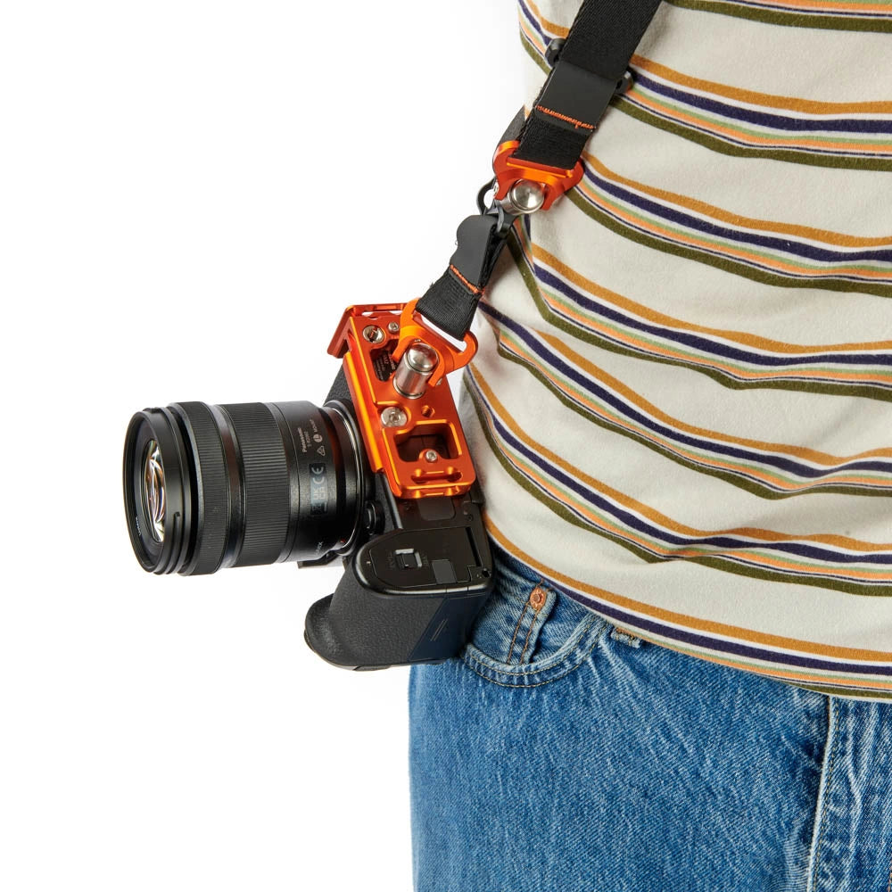 An orange camera rig mount holding a black camera body and lens, worn crossbody on a person's hip against a white background.