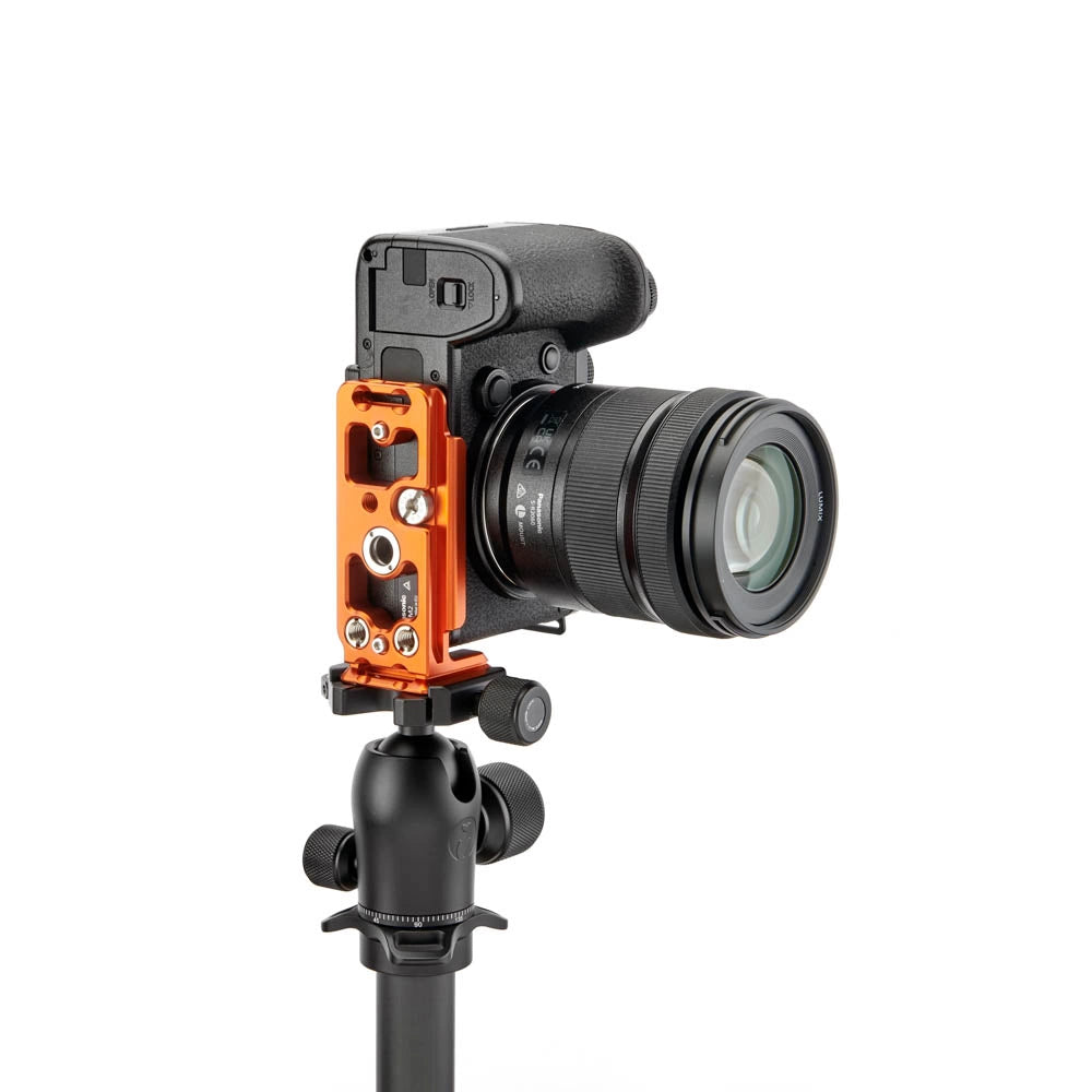 An orange camera accessory mount attached to a black DSLR camera with a lens, mounted on a black tripod, shown against a solid white background.