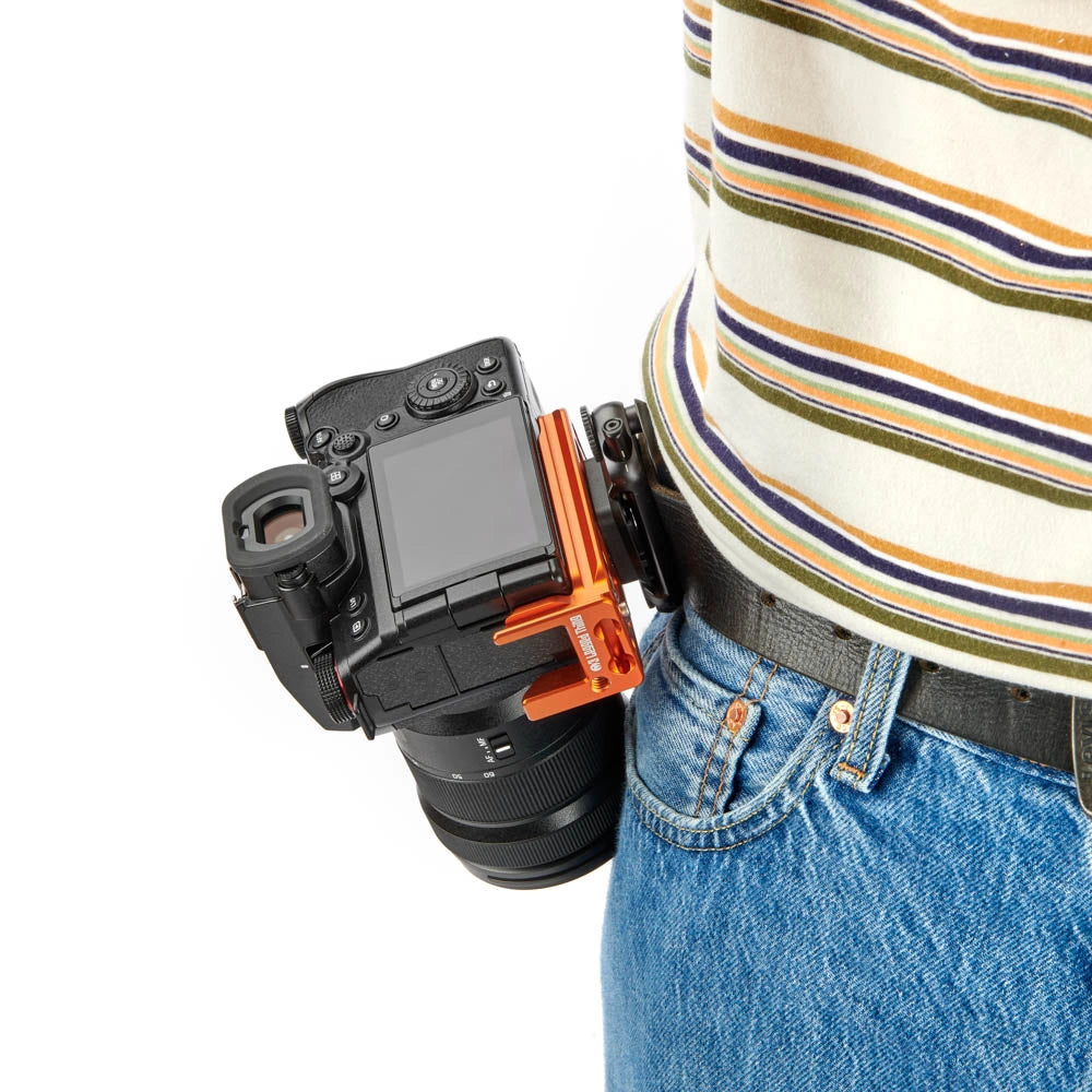 A black DSLR camera with an orange accessory mount attached to a black belt clip worn by a person in a striped shirt and blue jeans, shown against a plain white background.