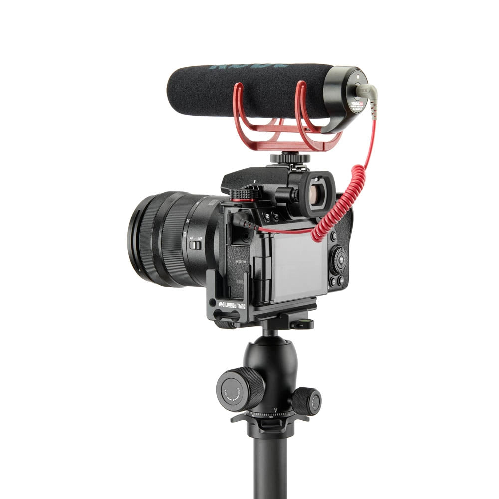 A black camera with a mounted lens, a red coiled cable, and a black shotgun microphone on a red stand, all attached to a black tripod, shown against a solid white background.