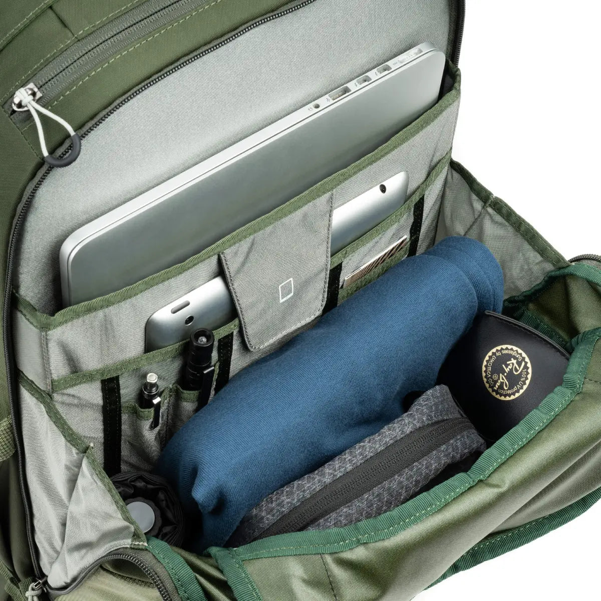 Green backpack with compartments containing electronic devices and a blue towel.