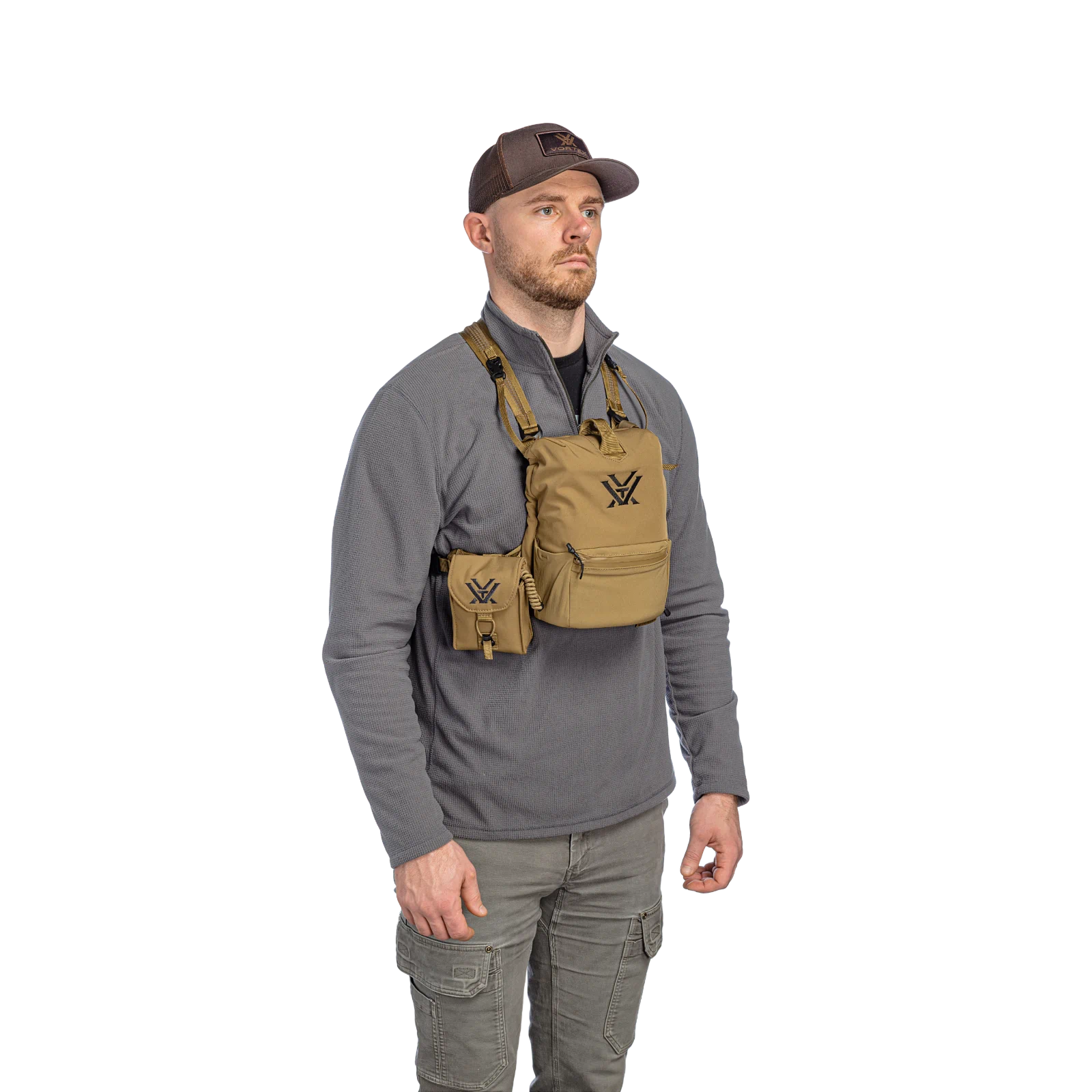 A man wearing a gray quarter-zip fleece, olive cargo pants, and a brown baseball cap models a tan harness with a matching pouch. The harness features a black embroidered logo and is shown against a solid white background.